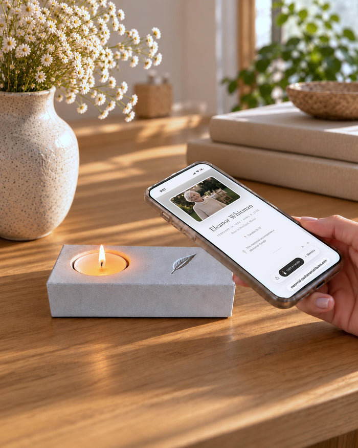 Concrete memorial candle holder with a lit tea light and an engraved feather. A hand rests a phone on the feather, opening an online memorial page.
