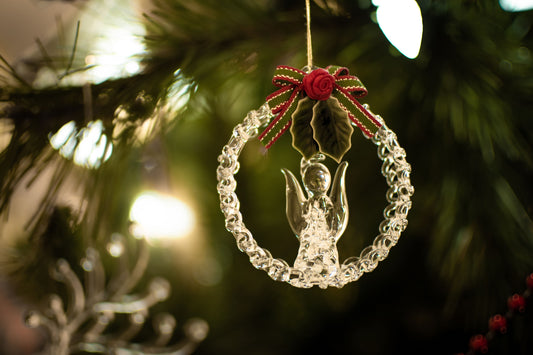 Clear glass angel ornament inside a glass wreath with a red ribbon and holly, hanging on a softly lit Christmas tree