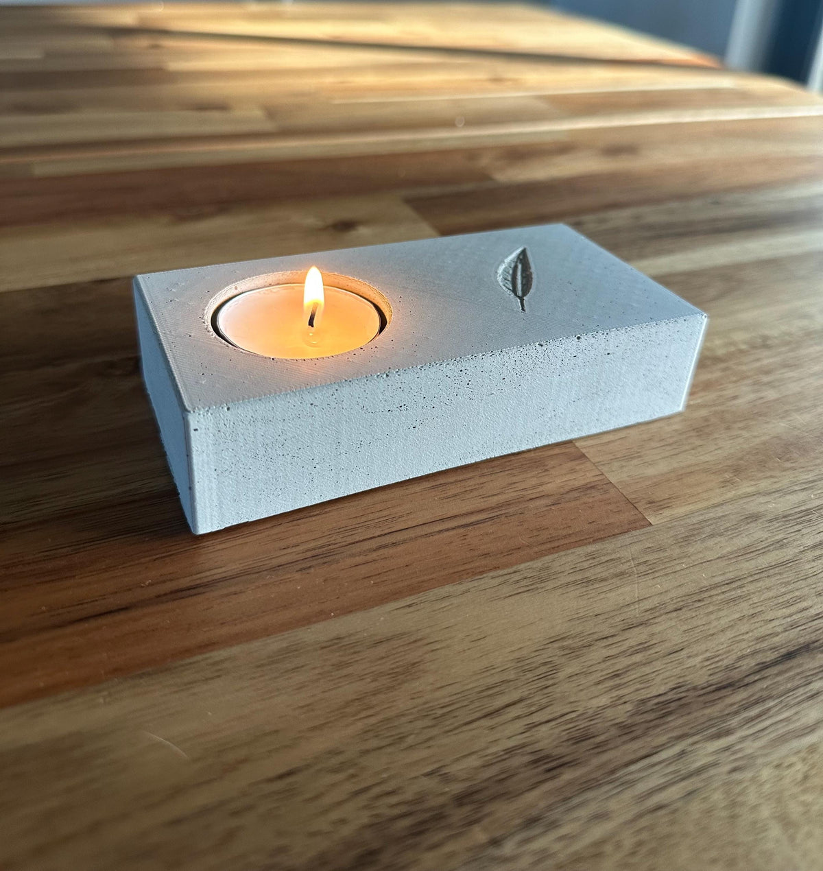 Unedited photo of the handmade concrete memorial candle holder with a lit tea light and engraved feather, on a wooden table.